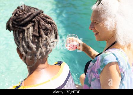Verschiedene ältere Freundinnen sitzen am Poolrand in gestreiftem Oberteil und schlürfen aus stiellosem Glas. Türkis, Wasser, Sonnenbrille, dr. Stockfoto