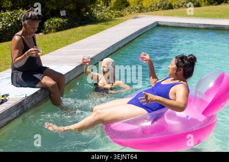 Drei verschiedene weibliche Freunde entspannen sich zu Hause am Pool in Badeanzügen und halten rosa Getränke auf dem Schwimmer. Wasser, Deck, Rasen, Sträucher, sonnig, Stockfoto