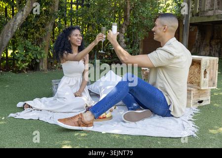 Ein Paar in weißem trägerlosem Kleid und Jeans, die Champagnerflöten auf einer Picknickdecke im Garten anschlagen. Romantisch, Garten, Toasten, Gläser, Kissen, shad Stockfoto