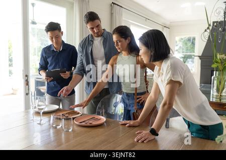Vielfältige Gruppe mit Tablets, die pinkfarbene Teller am Esstisch in einem modernen Zuhause anordnen. Zusammenarbeit, Planung, Diskussion, Tageslicht, openplan, Stockfoto