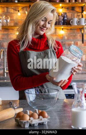 Frau mit grauem, rotem Pullover mit Schürze, der Mehlbecher hält und Teig in der Küche mit Schüssel-Eiern vorbereitet. Homebaker, gemütlich, rustikal, warm Stockfoto