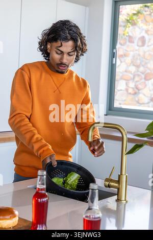 Indischer Mann mit orangefarbenem Sweatshirt, der Salat in schwarzem Sieb am Waschbecken hält. Greens, Sieb, Küche, modern Stockfoto