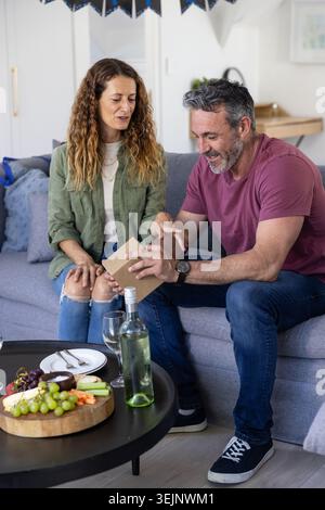 Ein Paar sitzt auf einem grauen Sofa, trägt Jeans und Hemden und hält eine kleine Karte mit Wurst und Wein. Wohnzimmer, minimalistisch, gemütlich Stockfoto