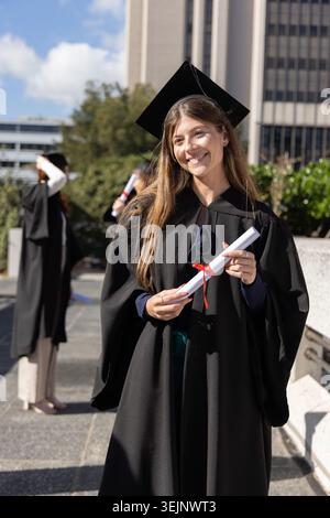 Weibliche Abiturin steht auf dem Campus plaza mit Mütze und Kleid, mit dem Diplom und dem lächelnden roten Band. Student, Universität, Zeremonie Stockfoto