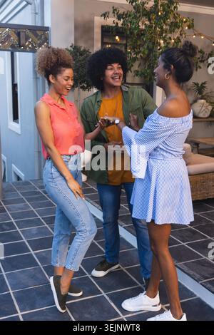 Verschiedene Freunde plaudern und lachen auf der gefliesten Terrasse unter Lichtern, in Korallenoberteil und gestreiftem Kleid. Im Freien, Terrasse, Treffen, Stockfoto