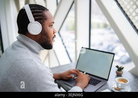 Junger erwachsener Mann, der Kopfhörer trägt, der einen Laptop benutzt, der aus der Ferne arbeitet Stockfoto