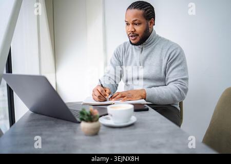 Erwachsener Mann schreibt Notizen in Notizbuch, mit Laptop-Computer Stockfoto