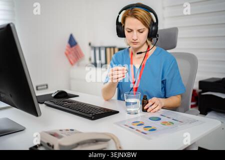 Krankenschwester in Peelings und Kopfhörern, die flüssige Medikamente am Schreibtisch einnehmen Stockfoto