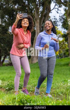Verschiedene Freundinnen laufen im Park in Sportbekleidung und machen Selfie mit Telefon und wiederverwendbarer Flasche. Im Freien, Grün, Bäume, fi Stockfoto