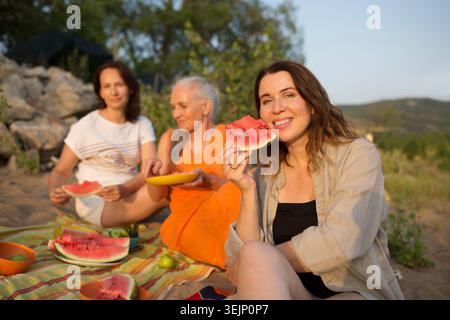 Drei Freundinnen mittleren Alters essen Wassermelone, während sie am Ufer beim Picknick sitzen Stockfoto