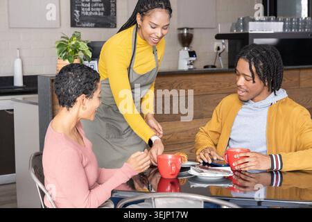 Afroamerikanerin Serviererin in gelber Schürze, die Gebäck auf schwarzen Kaffeetisch mit roten Tassen legt. Barista, Patrons, Espresso, Stockfoto