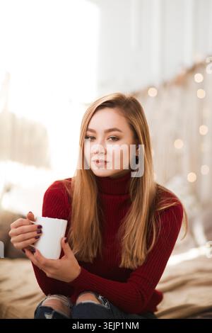 Glückliche junge Frau trinkt Kaffee oder Tee in der Nähe des Weihnachtsbaums. weihnachtskonzept am Morgen. Hygge-Konzept. Stockfoto