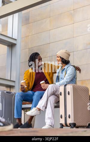 Verschiedene Paare sitzen auf der Bank und halten Kaffeetassen in der Bahnhofshalle mit beigefarbenem Koffer. Reise, Gepäck, Rucksack, Termina Stockfoto
