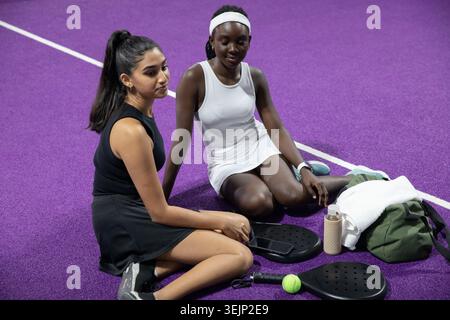 Verschiedene weibliche Mannschaftskameraden in sportlichen Kleidern sitzen auf dem purpurnen Platz mit Paddeln und gelbem Ball. Sportbekleidung, Schläger, Arena, Pos. Stockfoto