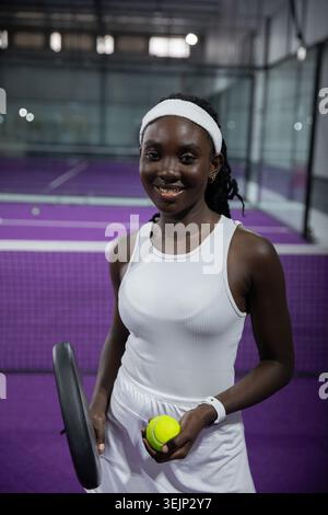 Afroamerikanerin im mittleren Erwachsenenalter, die auf einem violetten Court steht und weiß trägt, Schläger und Ball hält. Tennis, Sportler, Fitness, Focu Stockfoto