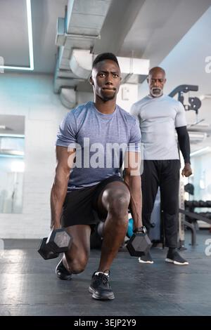 Leitender afroamerikanischer Trainer und männlicher Trainee, der einen gewichteten Ausfallschritt mit Hexen-Kurzhanteln im Fitnessstudio macht. Konzentration, Kraft, Fitness, Mment Stockfoto