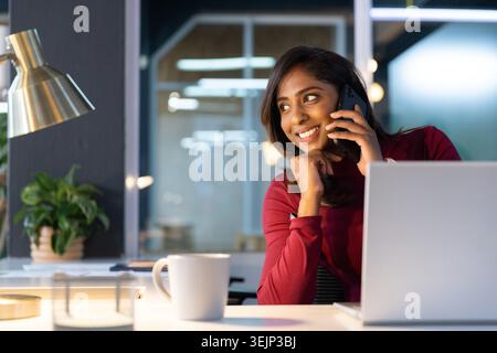Indische Frau sitzt am Schreibtisch im Büro, hält Smartphone mit offenem Laptop und trägt rotes Oberteil. Professioneller, moderner Arbeitsbereich Stockfoto