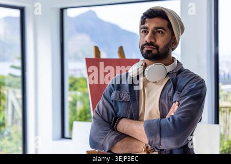 Indischer Mann mit Mütze und Kopfhörern, die an der Staffelei stehen, mit roter Leinwand und Fenstern, Kopierraum. Studio, Malerei, Tageslicht, Stockfoto