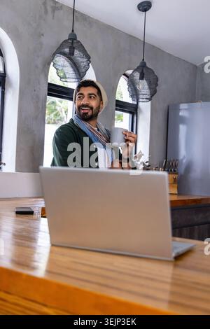 Indischer Mann, der hinter einer Holztheke in der modernen Küche steht und einen grauen Becher hält und einen Laptop benutzt. Person, Fernbedienung, freiberuflich, Studio Stockfoto