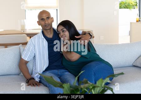 Diverse Paare sitzen auf grauem Sofa, Mann umarmen Frau, Pflanzen im Vordergrund, Holztisch sichtbar. Intimität, Gesellschaft, modern, Stockfoto