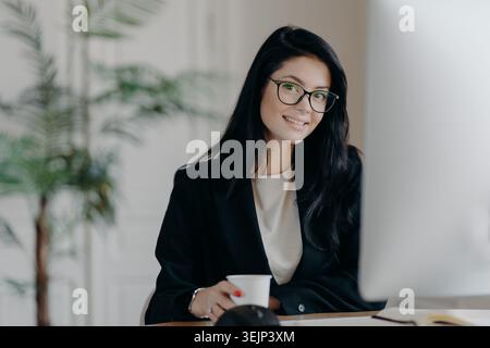 Lächelnde, zufriedene Ehefrau, die mit Forschungsarbeiten beschäftigt ist, posiert auf dem Schreibtisch vor dem Computermonitor, trinkt Kaffee gekleidet Stockfoto