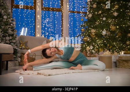 Frau, die Dehnübungen macht und Yoga-Fitness-Übungen im Weihnachtsinnenraum zu Hause macht. Fitness zu Hause, Aktivkleidung. Gesund und Stockfoto
