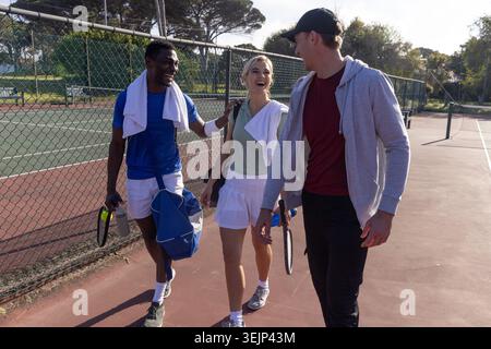 Verschiedene Freunde laufen auf dem Park Tennis Court in Tenniskleidung mit Schlägern, Seesack und Handtüchern. Trio, Lifestyle, realistisch, Stockfoto
