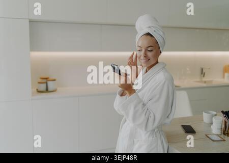 Glückliche gesunde Frau im Bademantel Anwendung Hautpflegeprodukt auf Gesicht nach dem Duschen oder Baden zu Hause Stockfoto