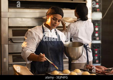 Bäckerinnen in Kochjacken und Haarnetz, die Teig mit Backbürste und Schüssel in der Bäckerei verglasten. Brotbackherstellung, Teamarbeit, Färbung Stockfoto
