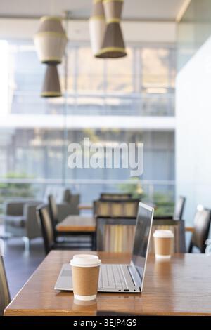 Offener silberner Laptop, der auf einem Holztisch mit Kaffeetasse aus Papier in der Nähe einer Glaswand sitzt. Coworking, Café, modern, ruhig, Tageslicht, min Stockfoto