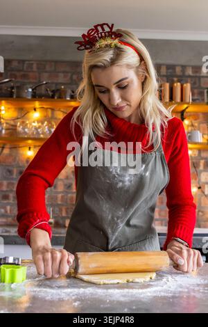 Frau in rotem Pullover in grauer Schürze rollender Teig mit hölzernem Rollstift an der Küchentheke. Bäcker, Zuhause, Mehl, gemütlich, Ziegelstein, sh Stockfoto