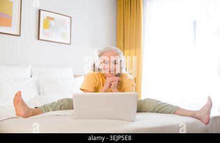Ältere Frau mit Kopfhörern streckt sich morgens mit Laptop auf einem Bett im Schlafzimmer. Gesunder Lebensstil. Online-Sportarten auf VI Stockfoto