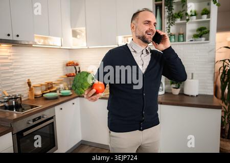 Mann, der am Telefon spricht und Gemüse in der Küche hält, lächelnd Stockfoto