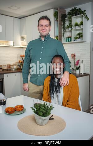 Interrassisches Paar posiert in der Küche, umarmt, lächelt in die Kamera Stockfoto