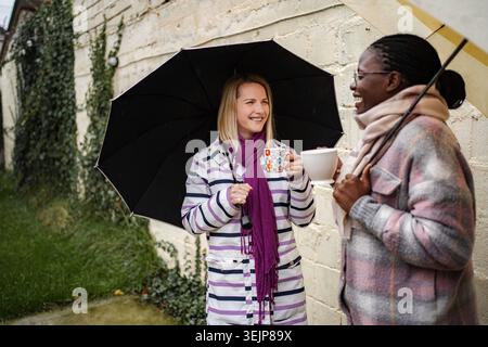 Zwei Frauen stehen unter Regenschirmen, lachen und trinken draußen Getränke Stockfoto