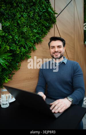 Junger erwachsener Mann lächelt und arbeitet an einem Laptop in einem modernen Café Stockfoto
