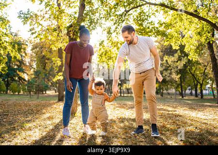 Eine fröhliche Familie genießt einen sonnigen Tag in einem Park, umgeben von Herbstlaub. Eltern halten ihr Kleinkind an den Händen, während sie es ermutigen, Schritte zu Unternehmen, cre Stockfoto
