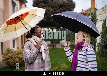 Zwei Frauen teilen sich einen fröhlichen Moment, lachen, während sie Regenschirme in den Händen halten Stockfoto