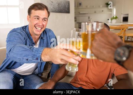 Verschiedene männliche Freunde, die Biergläser hochheben, zu Hause auf einem leichten Sofa mit Jeanshemd klirren, Uhr. Feiern, Toasten, Camara Stockfoto