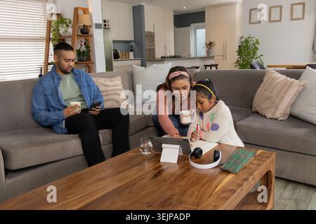 Vielfältige Familie, die sich zu Hause auf das Sofa lehnt und Kinder beim Schreiben am Tisch mit Tablet und Tassen begleitet. Gemütlich, Lounge, Eltern, Tochter, Col Stockfoto