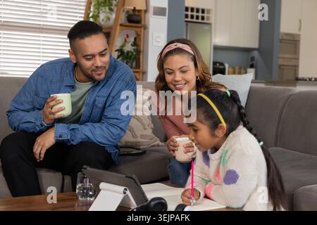 Vielfältige Familie, die zu Hause auf dem grauen Sofa sitzt und ein Tablet ansieht, während das Kind mit rosa Bleistift schreibt. Einheimisch, gemütlich, Innenraum, Modus Stockfoto