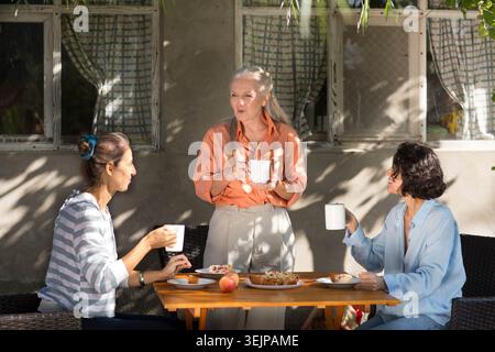 Drei Freundinnen aus der Mitte des Lebens trinken Tee und reden auf Stühlen in einem Sommergarten. Familientreffen. Teeparty Stockfoto
