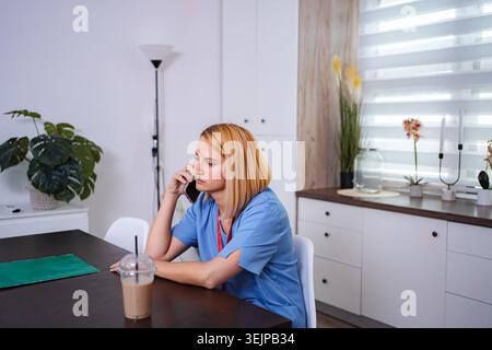 Eine Ärztin trägt blaue Peelings, die zu Hause in ihrer Küche telefoniert Stockfoto