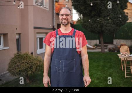 Ein lächelnder Mann auf der Schürze steht in einem lebendigen Hinterhof bei Sonnenuntergang Stockfoto