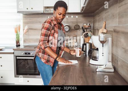 Junge Frau, Die Küchen-Zähler Reinigt, Während Sie Kaffeetasse Hält Stockfoto