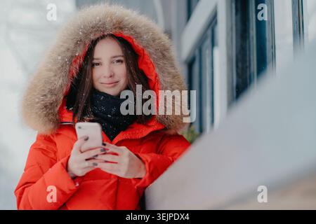 Außenaufnahme einer hübschen dunkelhaarigen Kaukasierin in winterlicher Straßenkleidung, hält modernes Handy, liest empfangene Nachricht, su Stockfoto