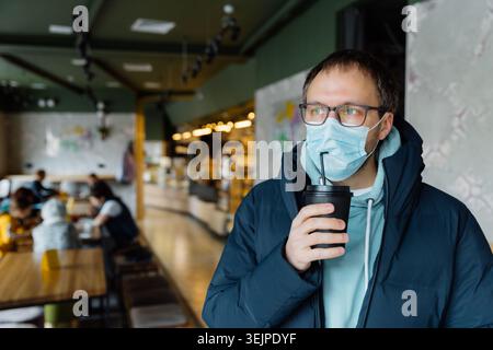 Innenaufnahme des ernsthaften nachdenklichen Mannes trinkt Kaffee zum Mitnehmen, trägt Schutzmaske, transparente Gläser, Jacke, posiert in der Menge Stockfoto