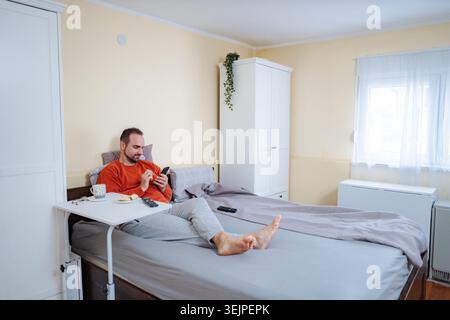 Mann, der sich im Bett entspannt und auf dem Smartphone beim Frühstück scrollt Stockfoto