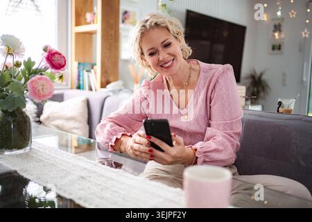 Frau lächelt, während sie das Telefon im Wohnzimmer benutzt Stockfoto