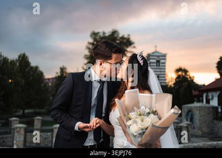 Interrassisches Hochzeitspaar küsst sich mit Blumenstrauß bei Sonnenuntergang Stockfoto